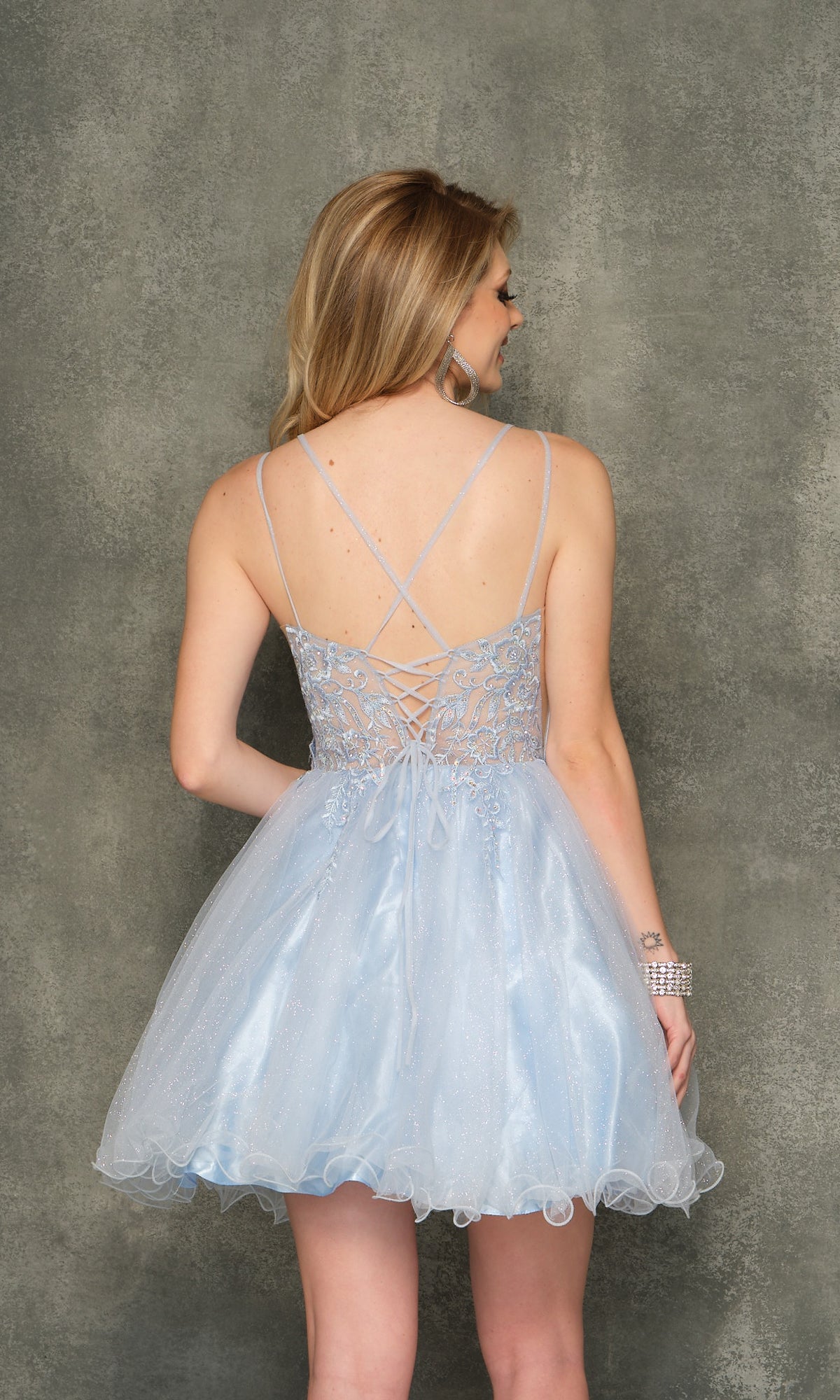 A woman wearing the Sheer-Bodice Ice Blue Short Homecoming Dress 10844, featuring a lace-up back, stands facing a textured gray wall.
